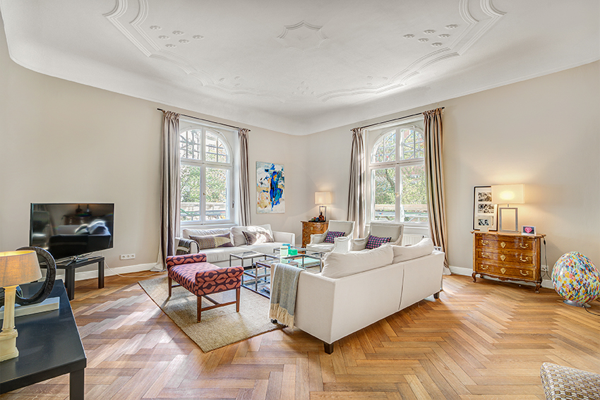 Ein geräumiges, helles Wohnzimmer mit großen Fenstern, Holzböden im Fischgrätmuster, zwei weißen Sofas, einem gemusterten Sessel, einem gläsernen Couchtisch, einem Fernseher, Kunstwerken an den Wänden und eleganten Deckendetails.