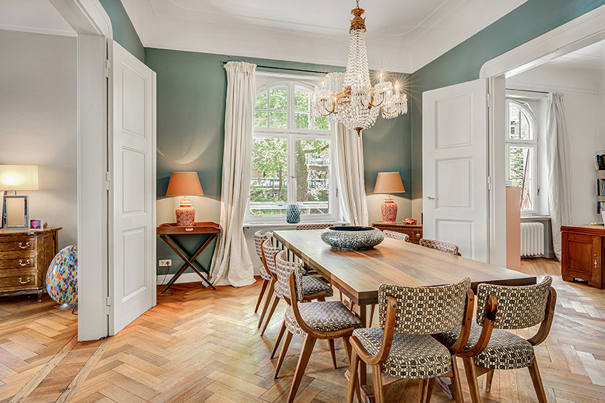 Ein stilvolles Esszimmer mit Holzböden, einem langen Holztisch, gemusterten Stühlen, einem Kristallkronleuchter und großen Fenstern mit weißen Vorhängen, die natürliches Licht hereinlassen. Beistelltische mit Lampen stehen auf beiden Seiten des Fensters.