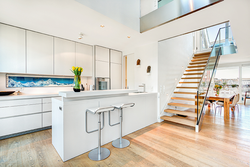 Moderne, offen gestaltete Küche mit weißen Schränken, einer eleganten Insel mit zwei grauen Barhockern, Holzfußboden und einer Schwebetreppe, die in ein höheres Stockwerk führt. Große Fenster lassen viel natürliches Licht herein.