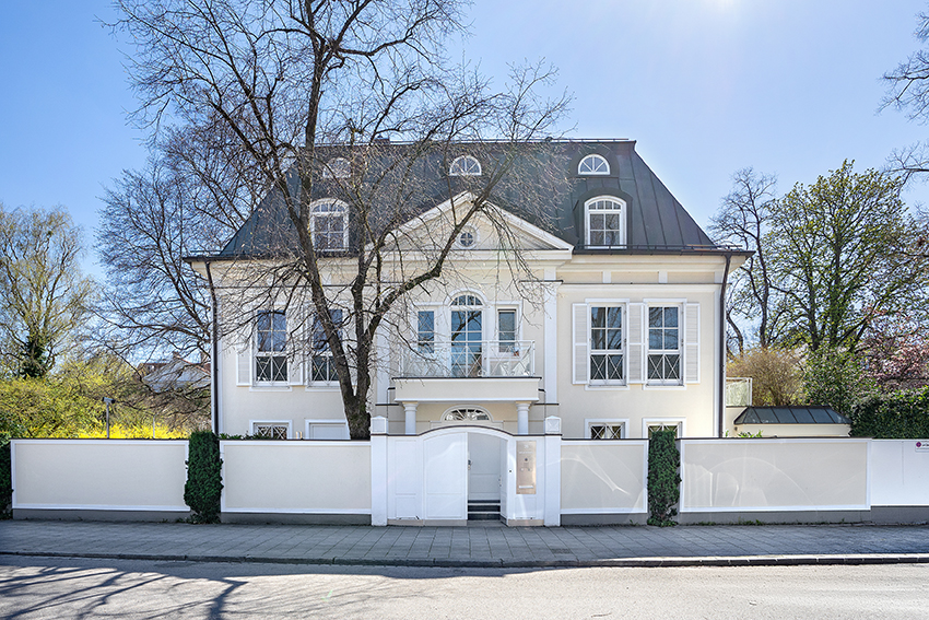 Ein großes, elegantes weißes Haus mit hohen Fenstern, einem dunklen Dach und einem weißen Eingangstor, umgeben von einer weißen Mauer und Bäumen unter einem klaren blauen Himmel.