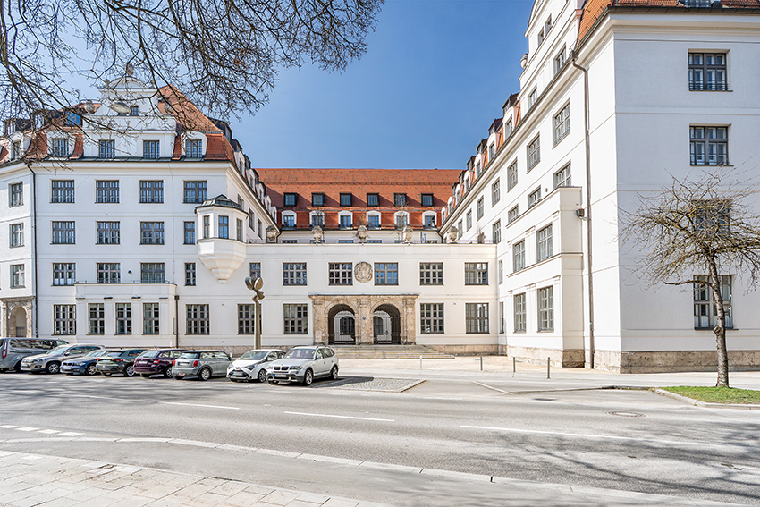 Ein großes, weißes historisches Gebäude mit rotem Ziegeldach, vielen Fenstern und einem bogenförmigen Eingang. An der Straße davor sind mehrere Autos geparkt, und an einem klaren, sonnigen Tag sind ein paar blattlose Bäume zu sehen.