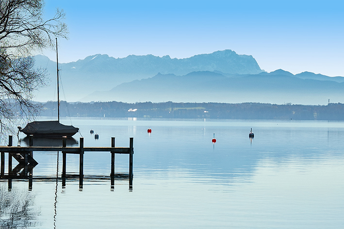 Ein ruhiger Ammersee mit einem Steg und einem überdachten Segelboot auf der linken Seite, mehreren schwimmenden Bojen und entfernten blauen Bergen unter einem klaren Himmel. Im Wasser spiegelt sich die ruhige Landschaft.