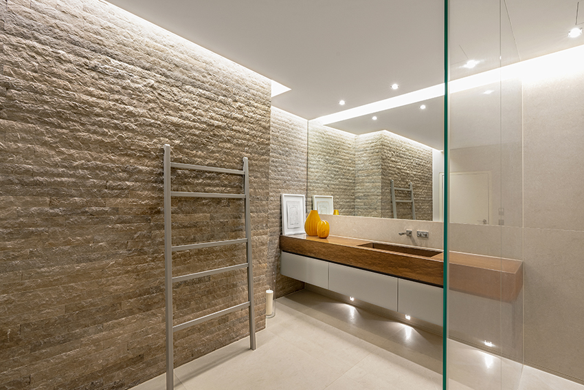 Modernes Badezimmer mit strukturierter Steinwand, großem Spiegel, hölzerner Arbeitsplatte mit weißen Schubladen, gelbem Vasen-Dekor, Handtuchleiter und gläserner Duschabtrennung unter versenkter Beleuchtung.