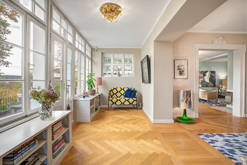 Heller, eleganter Wohnraum mit großen Fenstern, Parkettboden, Bücherregalen, einem gelb gemusterten Sessel, Zimmerpflanzen und stilvollem Dekor. Licht strömt herein und verleiht dem Raum ein luftiges, einladendes Gefühl.