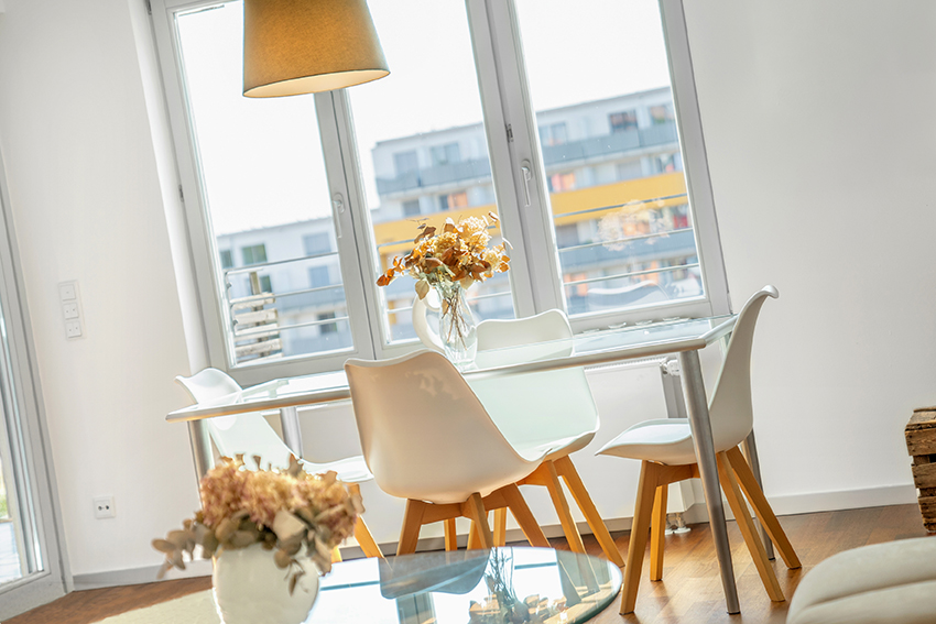 Ein moderner Essbereich mit einem Glastisch, vier weißen Stühlen mit Holzbeinen, einer Blumenvase auf dem Tisch, einer Hängelampe über dem Tisch und großen Fenstern, die natürliches Licht hereinlassen, während die Gebäude draußen sichtbar sind.