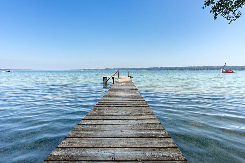 Ein hölzerner Steg erstreckt sich über ruhiges blaues Wasser bis zum Horizont unter einem klaren Himmel. Auf der rechten Seite ist ein einzelnes rotes Segelboot zu sehen und im Hintergrund ein entferntes, von Bäumen gesäumtes Ufer.